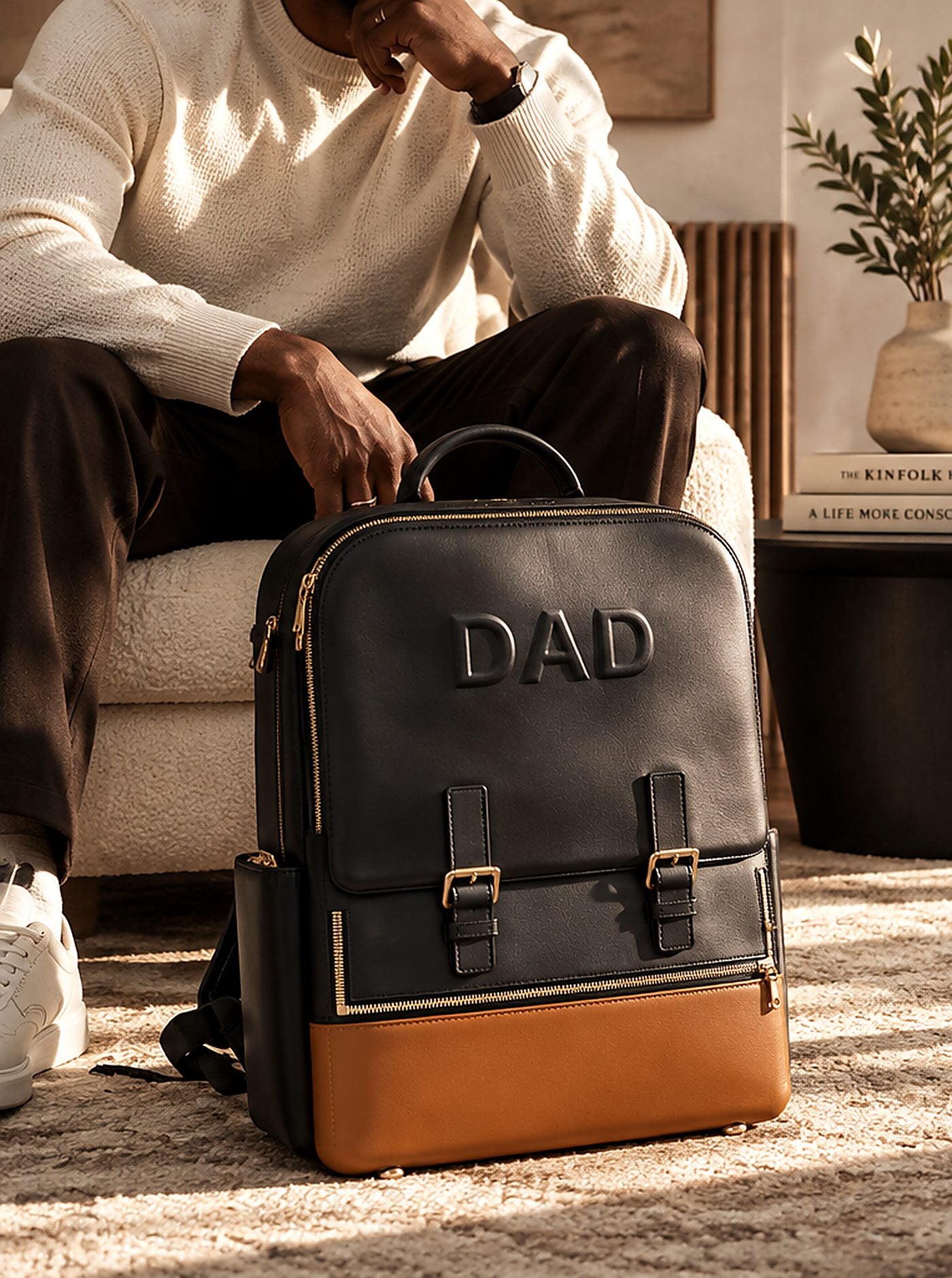 The DAD Backpack in black with a cognac base and two buckle straps, beside a father seated in a sunlit living room